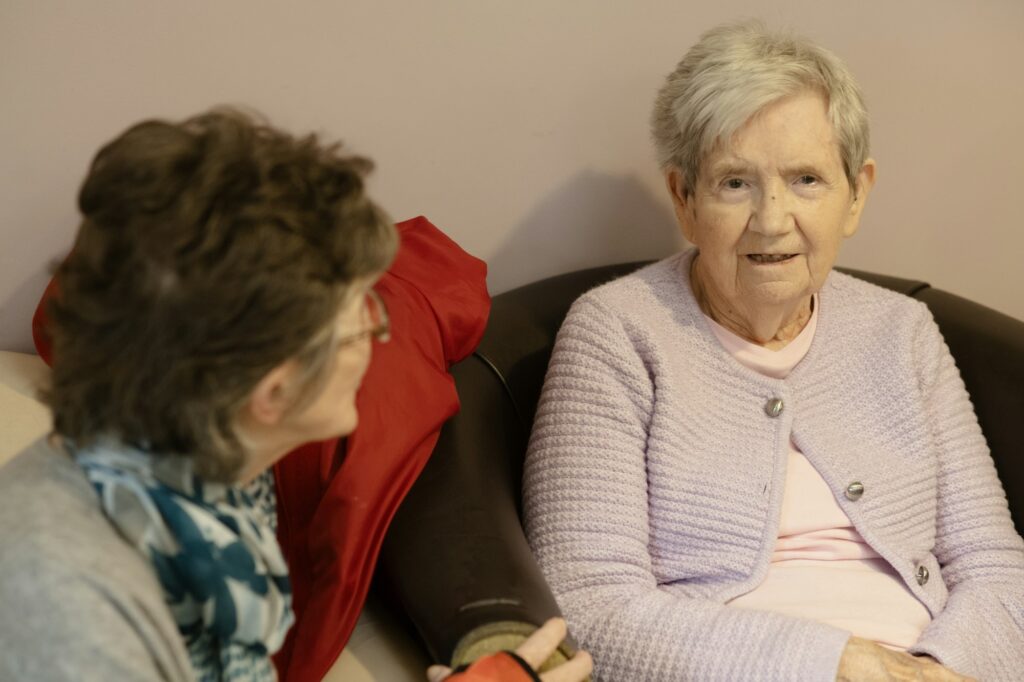 An older woman sits in a chair conversing with another woman seated nearby in a calm, well-lit consultation space.