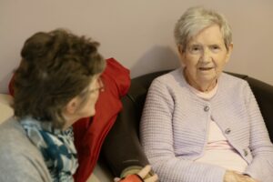 An older woman sits in a chair conversing with another woman seated nearby in a calm, well-lit consultation space.