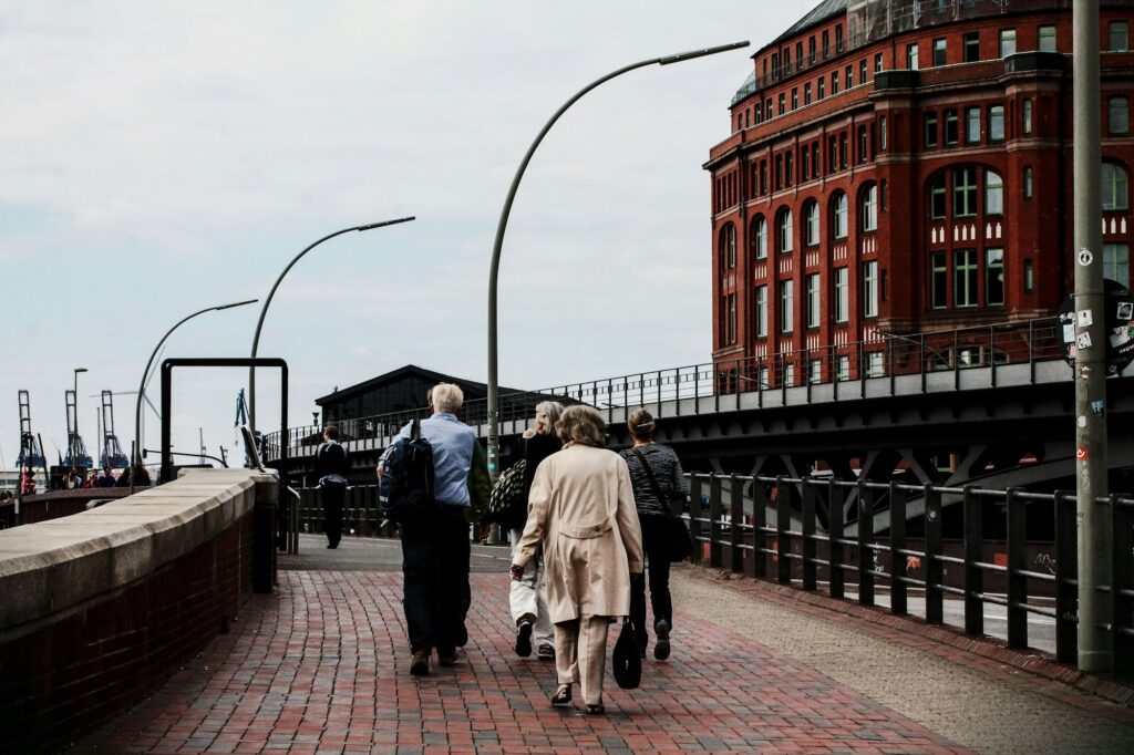 A group of older adults walks along a brick promenade beside a river with a historic red brick building in the background.